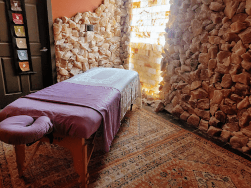 A serene massage room with a stone wall, a massage table covered in a purple cloth, and warm lighting.